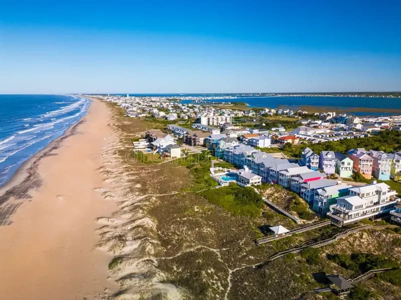 Aerial view of beachfront houses along a long sandy coastline with the ocean on one side and a bay on the other under a clear blue sky