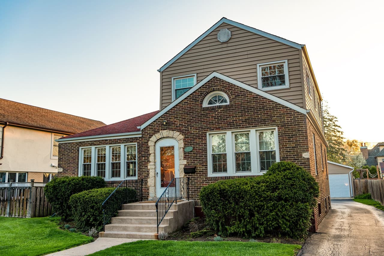 Two-story brick suburban house with beige siding on upper floor, front steps leading to a white door, surrounded by green bushes and lawn, under clear sky during sunset