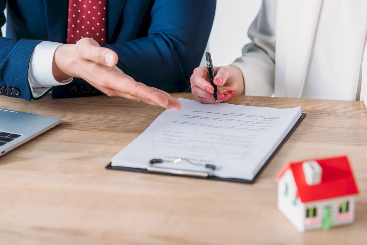 A businessman and a woman sitting at a table with a contract on a clipboard. The woman is signing the contract while the businessman gestures with his hand. A small model house is on the table, indicating a real estate or legal agreement.