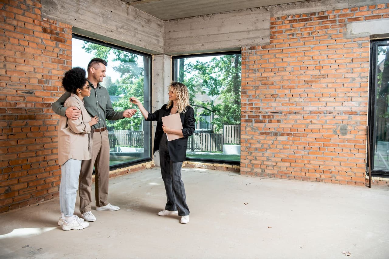 A female real estate agent talking to a couple inside an unfinished brick house with large windows showing greenery outside