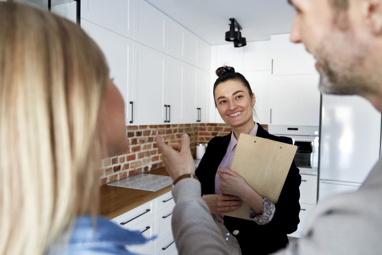 Female real estate agent holding clipboard and talking to a couple in a modern kitchen with white cabinets and brick backsplash