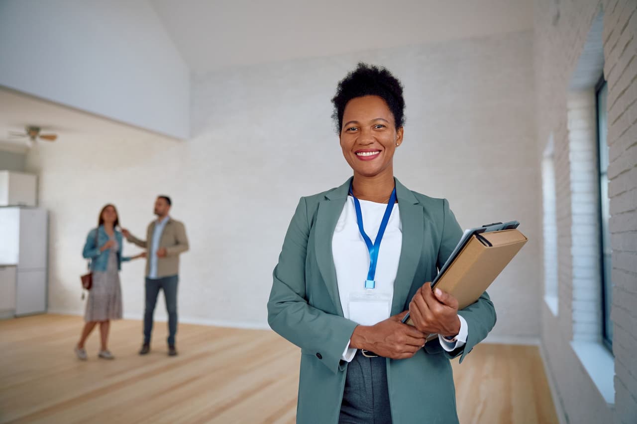 Portrait of a smiling female real estate agent holding a clipboard in an empty room with two people talking in the background