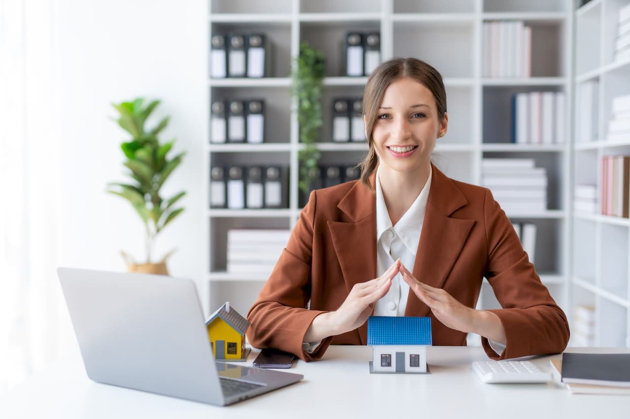 Portrait of a smiling female real estate agent sitting at a desk with a laptop and miniature house models, with a blurred office background featuring shelves and plants.