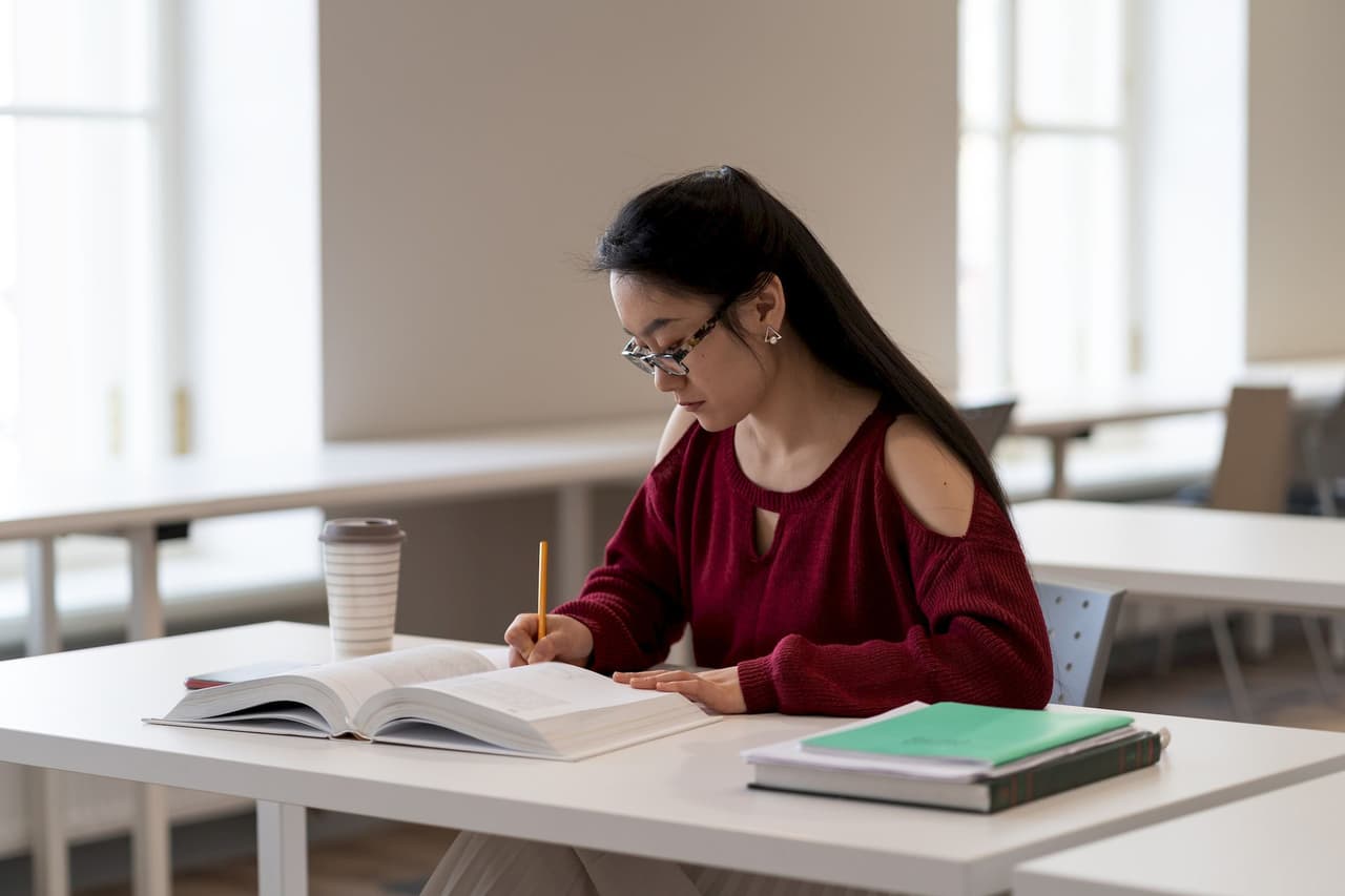 A female student with long dark hair and glasses is studying at a white table in a library, writing in a notebook with an open book and a coffee cup nearby.