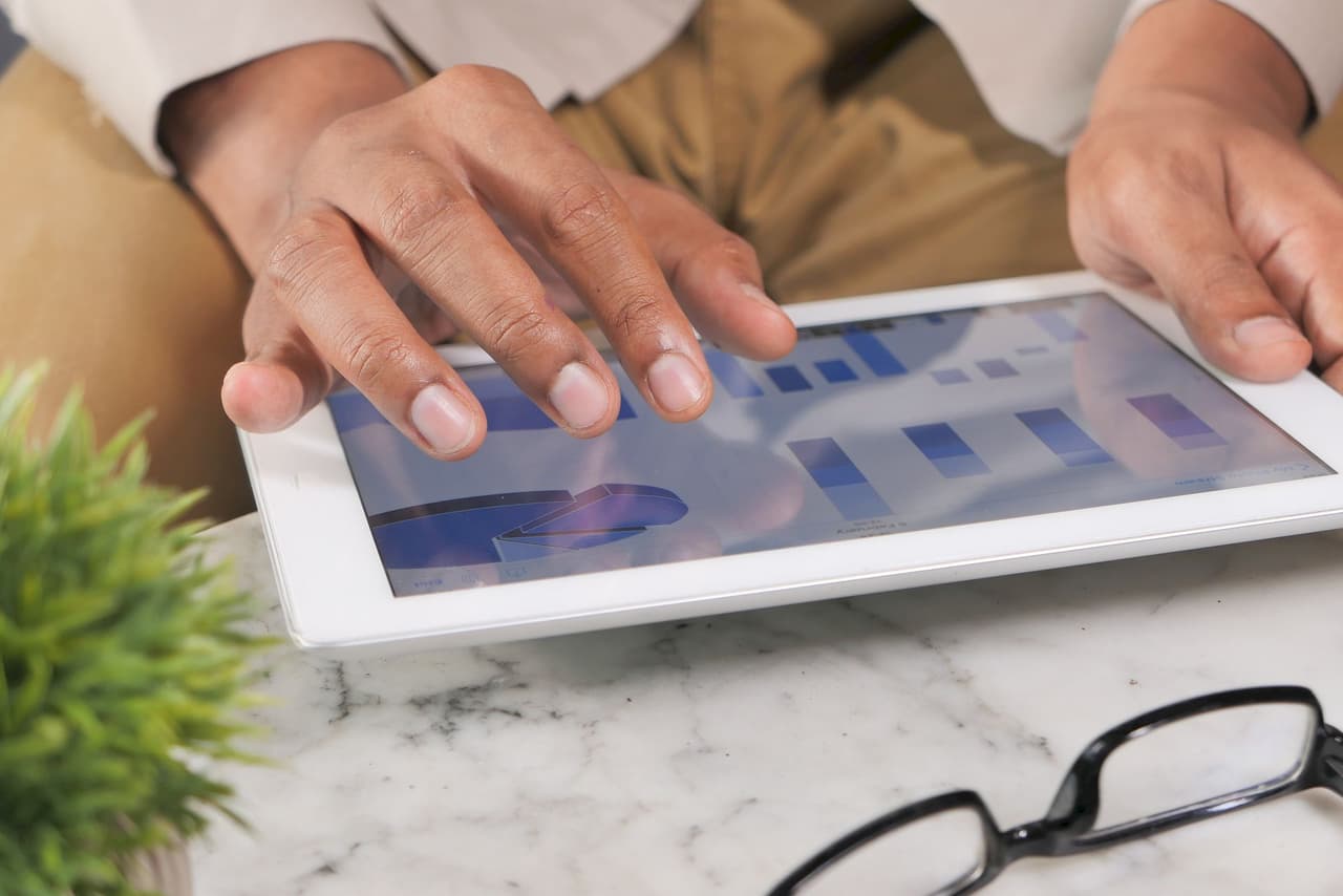 Close-up of male hands interacting with a tablet device on a marble surface, with a blurred background including a plant and eyeglasses