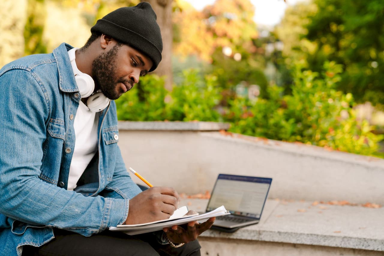 Young male student wearing a black beanie and denim jacket, sitting outdoors on concrete steps, writing notes on a clipboard with a laptop open beside him, surrounded by greenery and trees in the background.