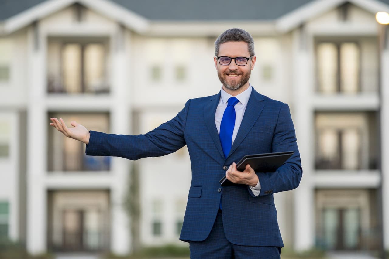 Smiling man in business suit holding tablet and gesturing with hand in front of blurred building background