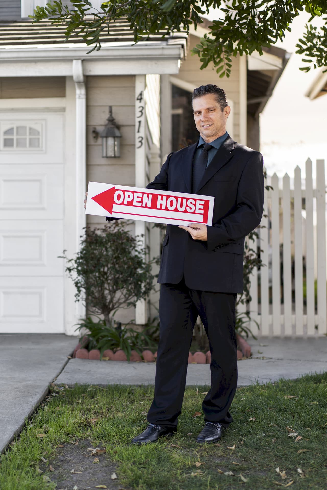 Man in black suit holding an open house sign in front of a residential home with a white picket fence and greenery