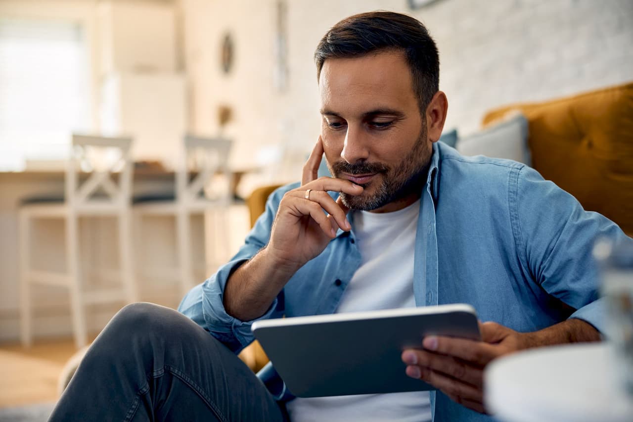 A man with short dark hair and beard sitting indoors, using a tablet with a blurred background of a cozy room.