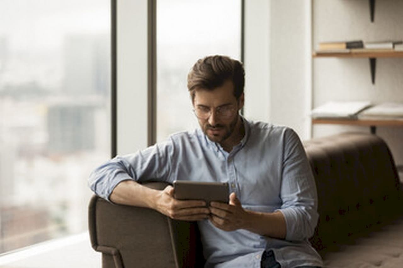 A man with glasses sitting on a couch indoors near a large window, focused on using a tablet device, with a blurred cityscape background.