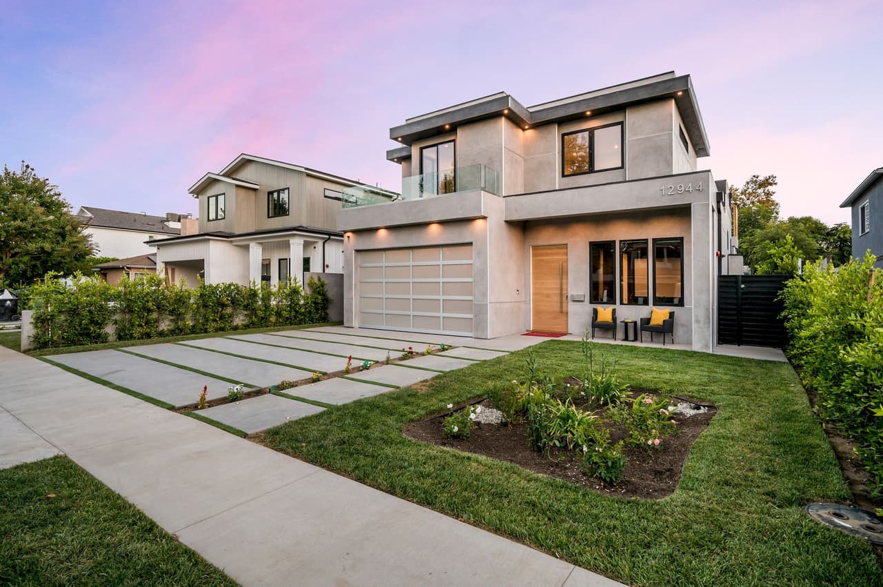 Modern suburban house exterior with a two-car garage, well-maintained lawn, and garden bed under a pink and purple evening sky