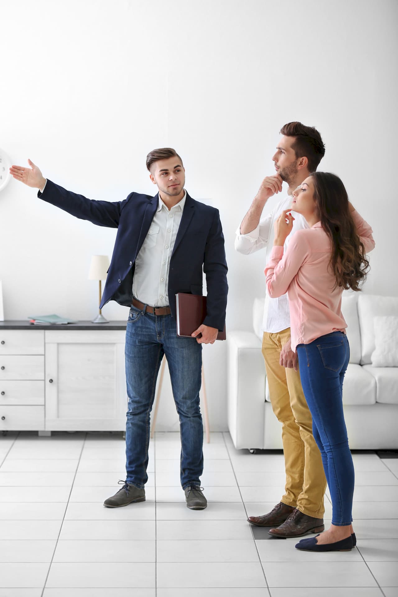 Real estate agent showing a young couple a property interior, discussing and pointing towards a window in a bright room