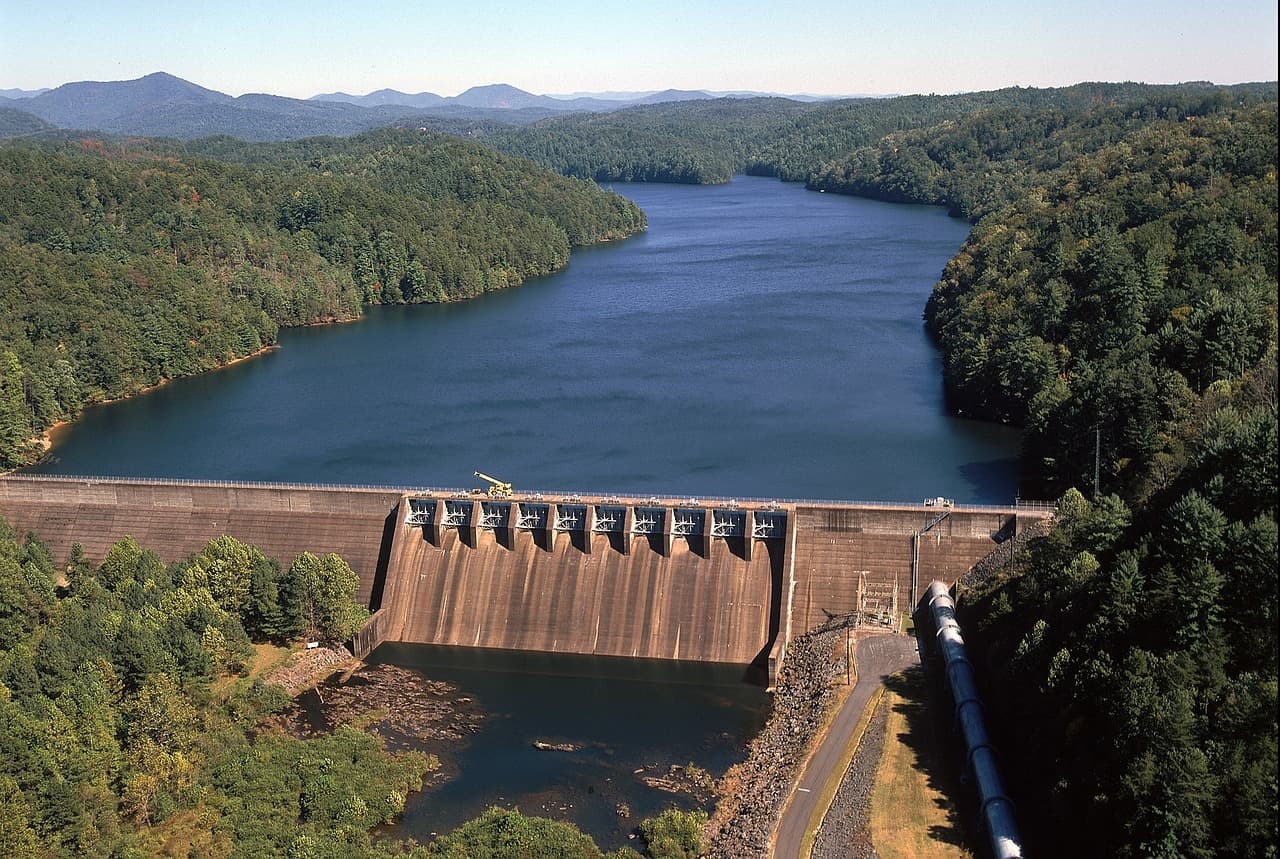 A large dam across a wide river surrounded by dense green forest with mountains in the background under a clear sky