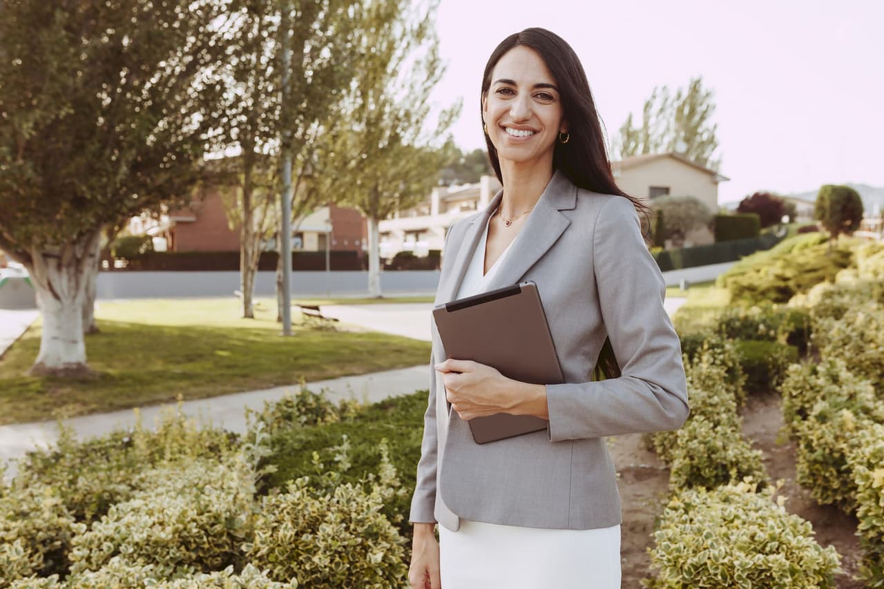 Portrait of a smiling woman in business attire holding a folder outdoors with greenery and trees in the background