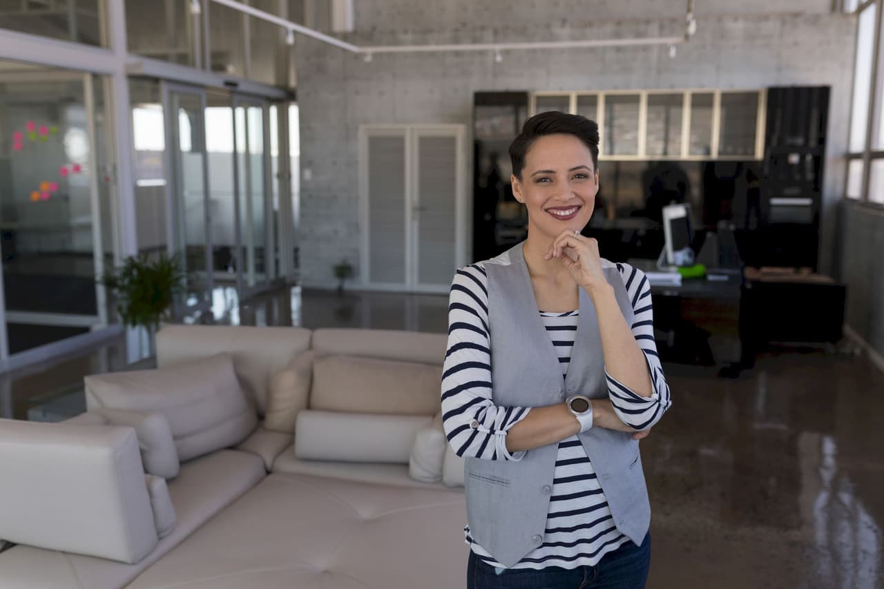 Smiling woman standing in a modern office with a blurred background, wearing a striped shirt and gray vest.
