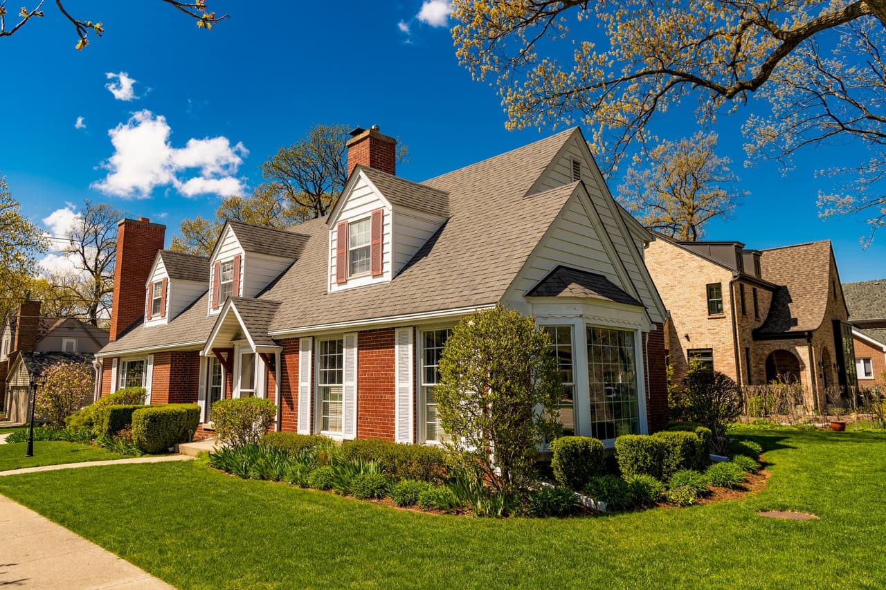 A suburban house with brick and white siding under a clear blue sky with some clouds, surrounded by green grass and trees in daytime.