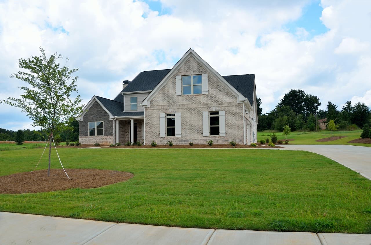 A suburban two-story house with a green lawn and a tree in the front yard under a partly cloudy sky