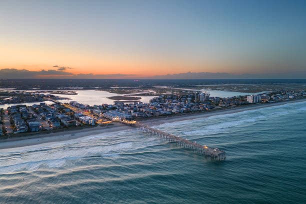 Aerial view of a coastal town with a pier extending into the ocean during sunset, showing calm waters and a colorful sky.