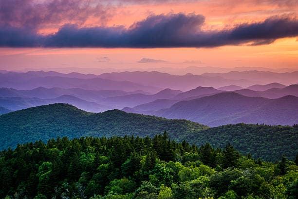 Scenic view of rolling mountain ridges covered in forest under a colorful sunset sky with layered clouds