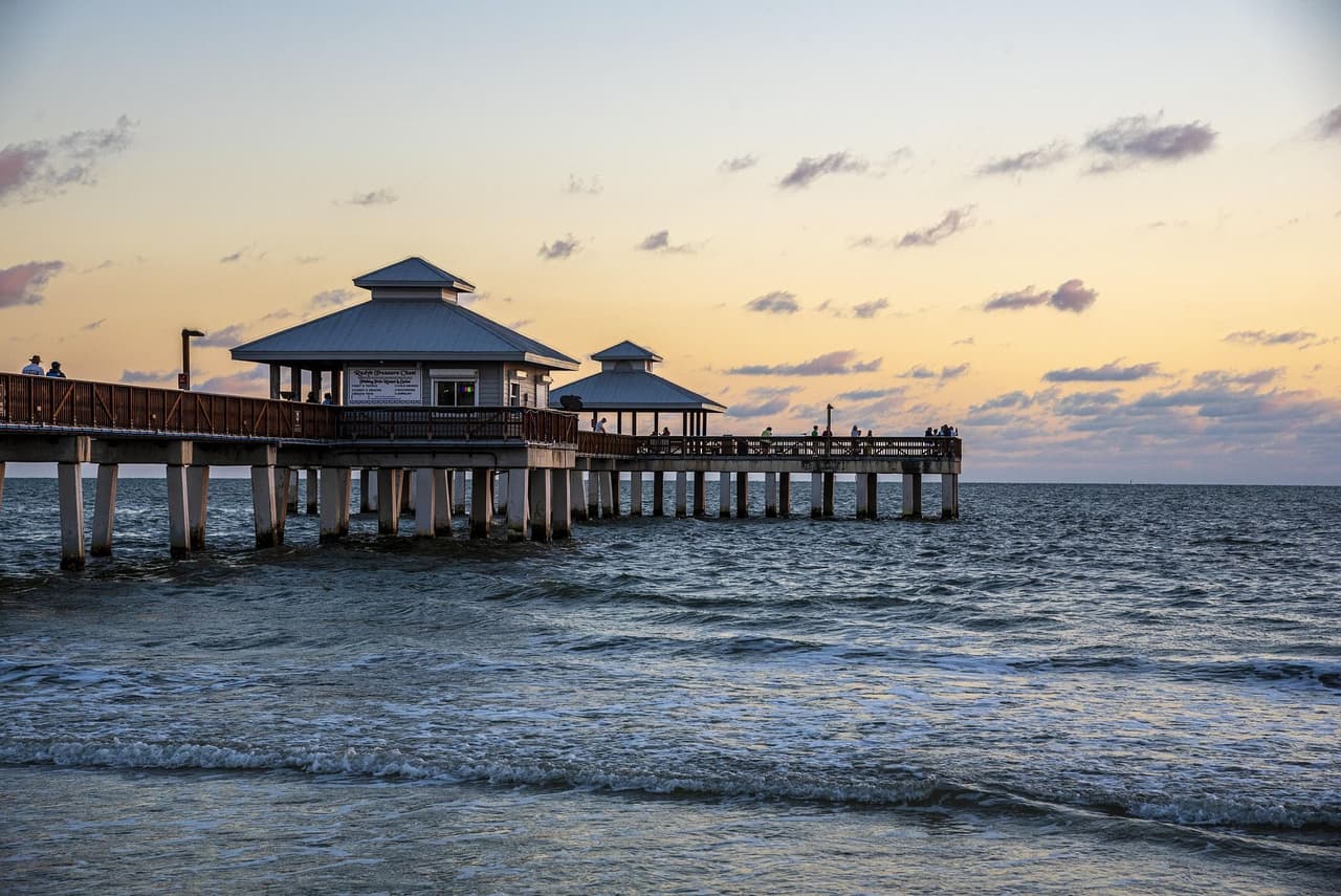 Wooden pier with small pavilion structures extending into ocean waves at sunset with people walking and standing on pier under a partly cloudy sky