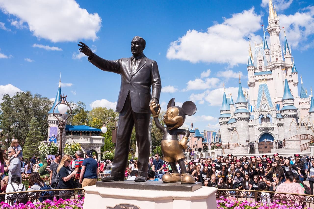 Statue of a male figure in a suit holding hands with Mickey Mouse in front of a large castle with a crowd of people in the background