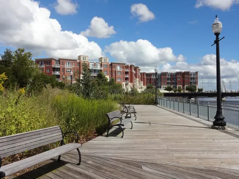 Wooden boardwalk with benches along waterfront, buildings in background under blue sky with clouds
