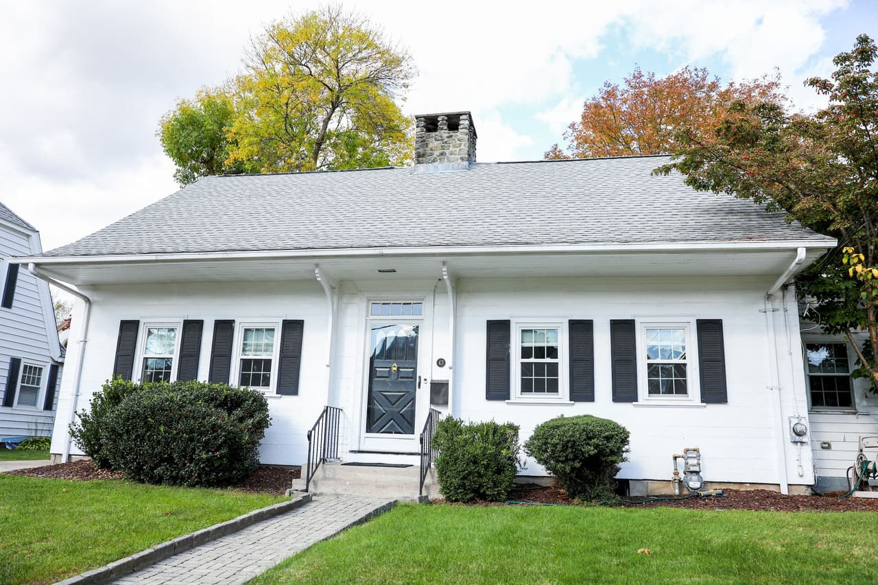 Front view of a white cottage house with black shutters, a gray roof, and a green lawn in the front yard with trees in the background