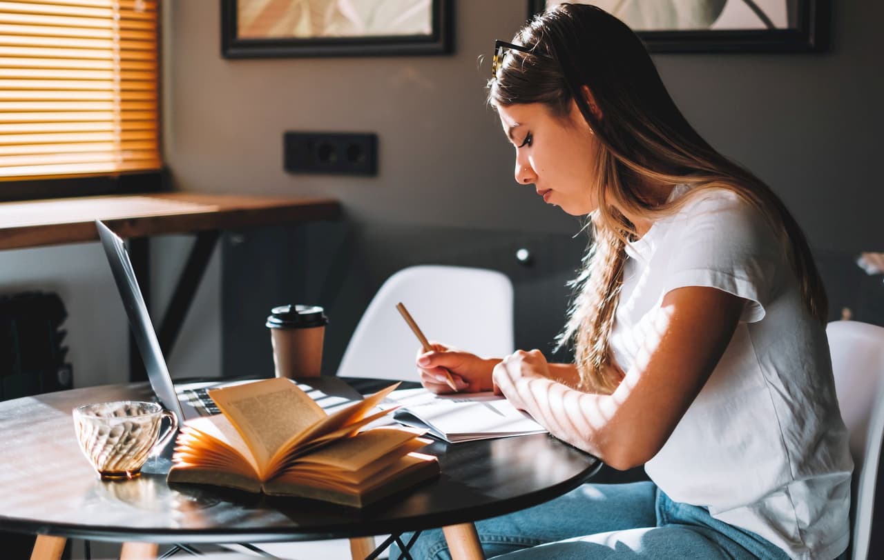 Young woman studying and writing in a notebook at a round table in a cafe with an open book, laptop, and coffee cup, natural light from window