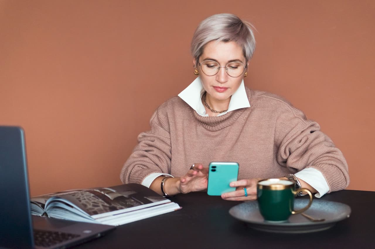 A woman with short gray hair and glasses is sitting at a table using a smartphone. On the table are an open book, a laptop, and a green cup with a gold handle. The background is a solid warm brown color.