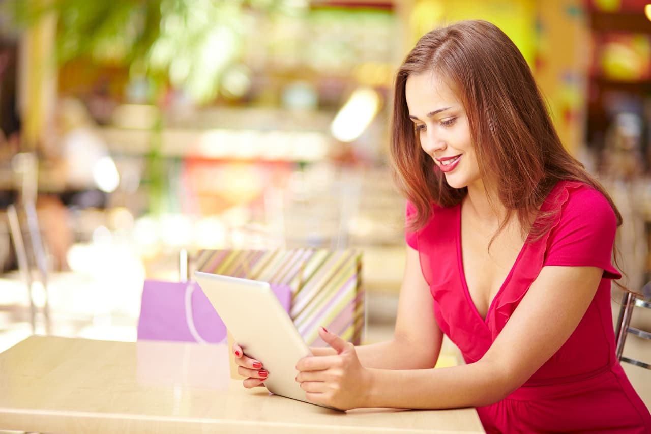 Portrait of a woman with brown hair wearing a red dress, sitting at a table in a cafe, using a tablet with a blurred background.