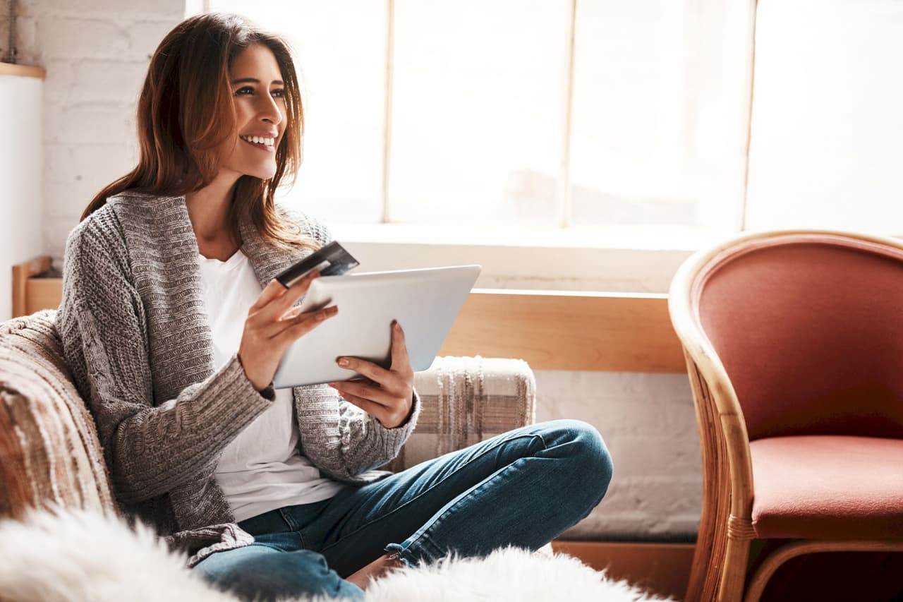 Smiling woman sitting on couch using tablet and holding credit card in bright room