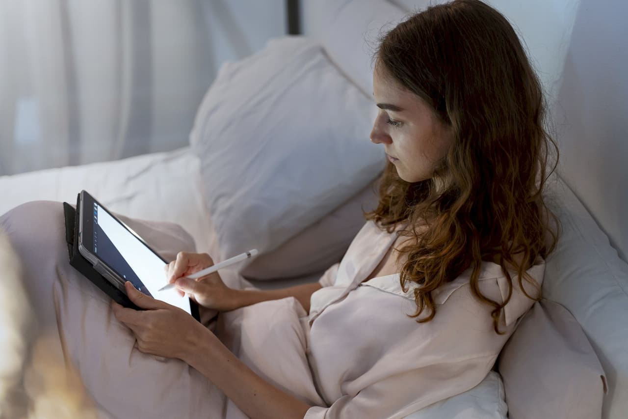 Young woman with long hair using a tablet with a stylus while sitting in bed with pillows, soft natural lighting
