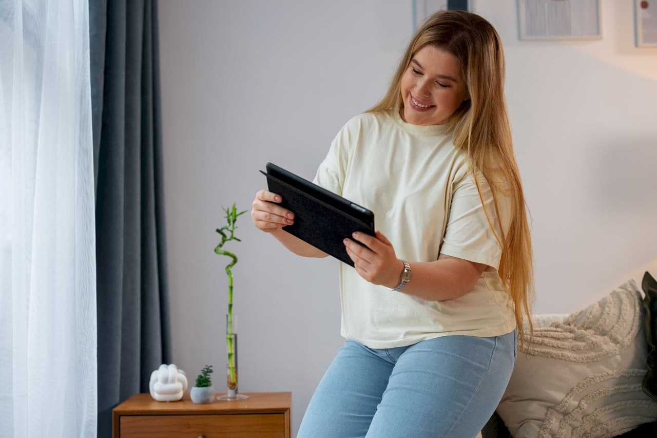 A young woman with long hair sitting indoors on a bed, smiling and using a tablet device in a casual setting with a small plant and curtain in the background.