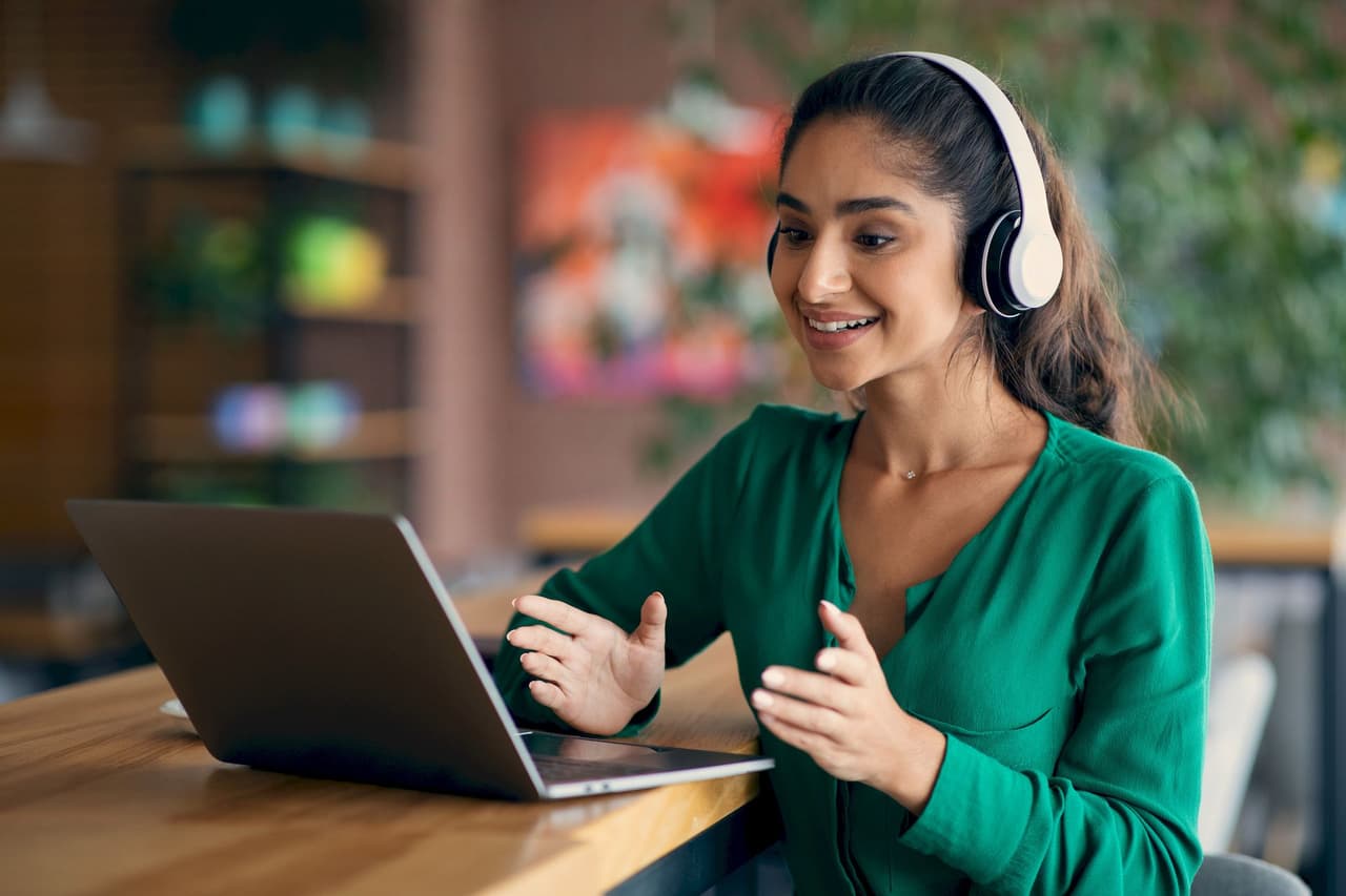 Young woman wearing headphones smiling and gesturing while having a video call on a laptop with a blurred background