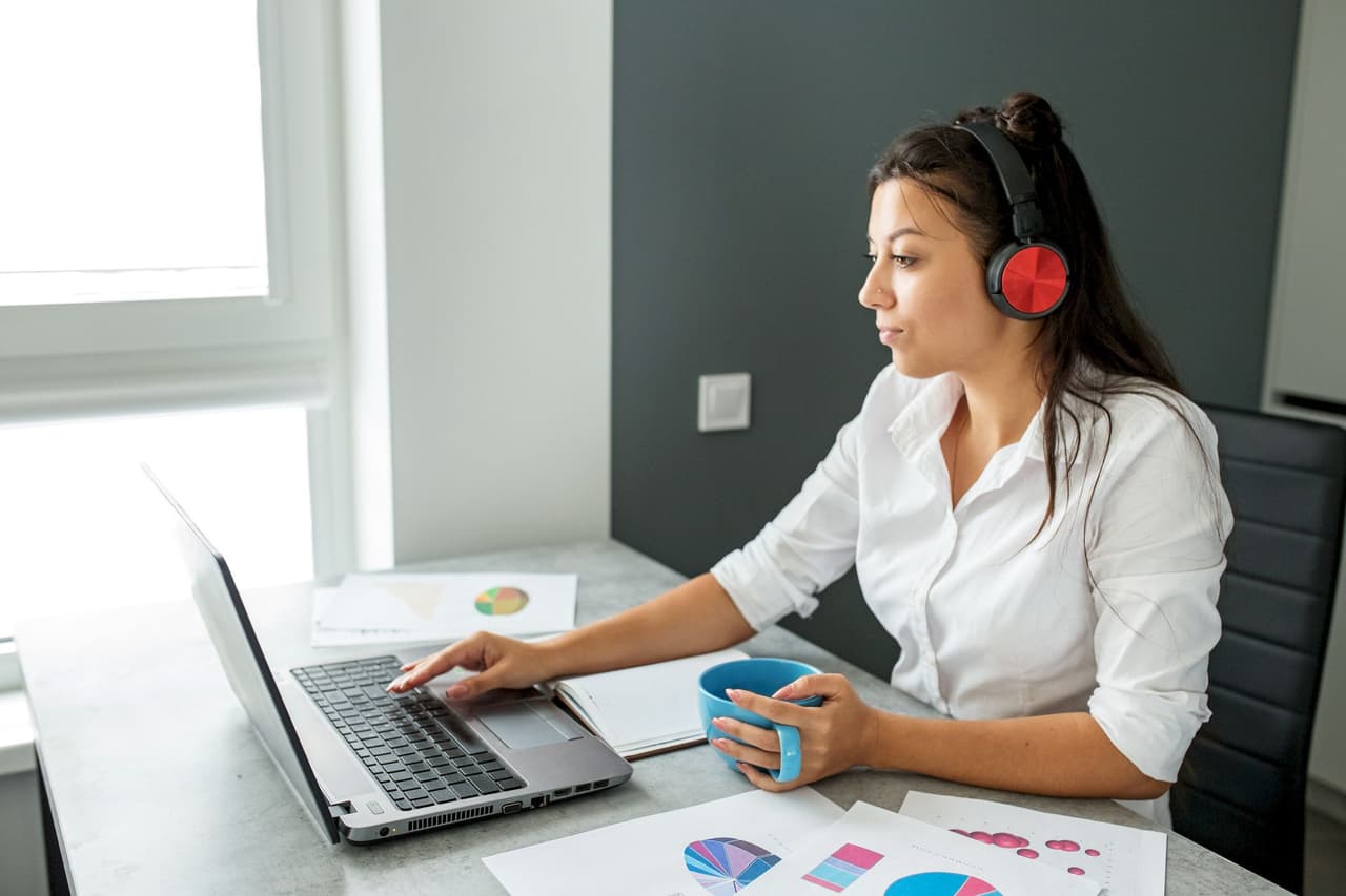 A woman wearing red headphones working on a laptop at a desk with charts and a blue mug, sitting in a modern office with natural light from a window