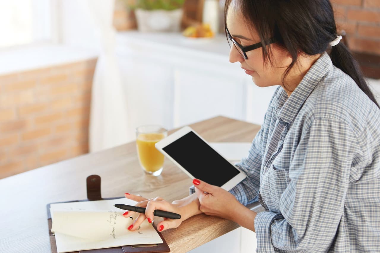 A woman with glasses writing notes in a notebook while holding a tablet, sitting at a wooden table with a glass of orange juice, with a blurred background.
