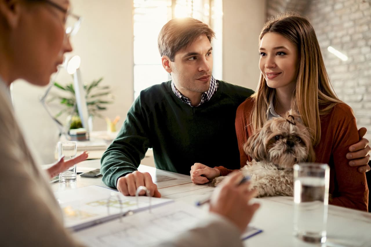 Young man and woman sitting at a desk with an insurance agent, woman holding a small dog, indoor office setting