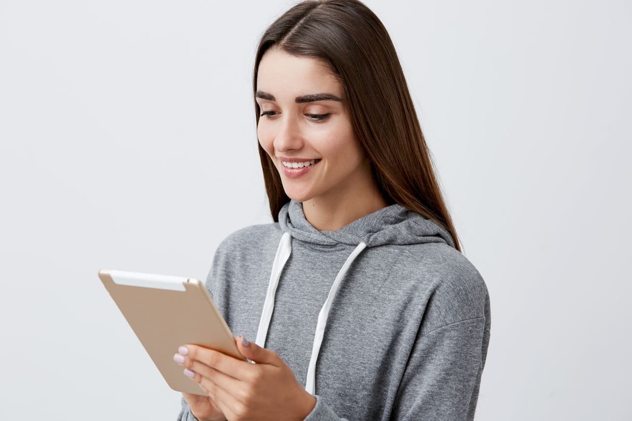 Portrait of a young woman in a gray hoodie smiling and looking at a tablet against a plain light gray background