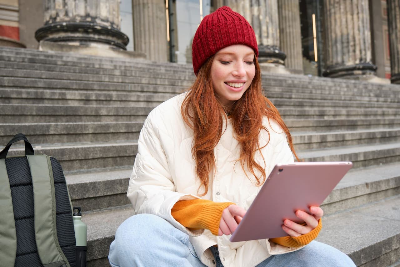 Young woman with red hair wearing a red beanie and white jacket sitting on stone steps outdoors, smiling and using a tablet, with a backpack beside her