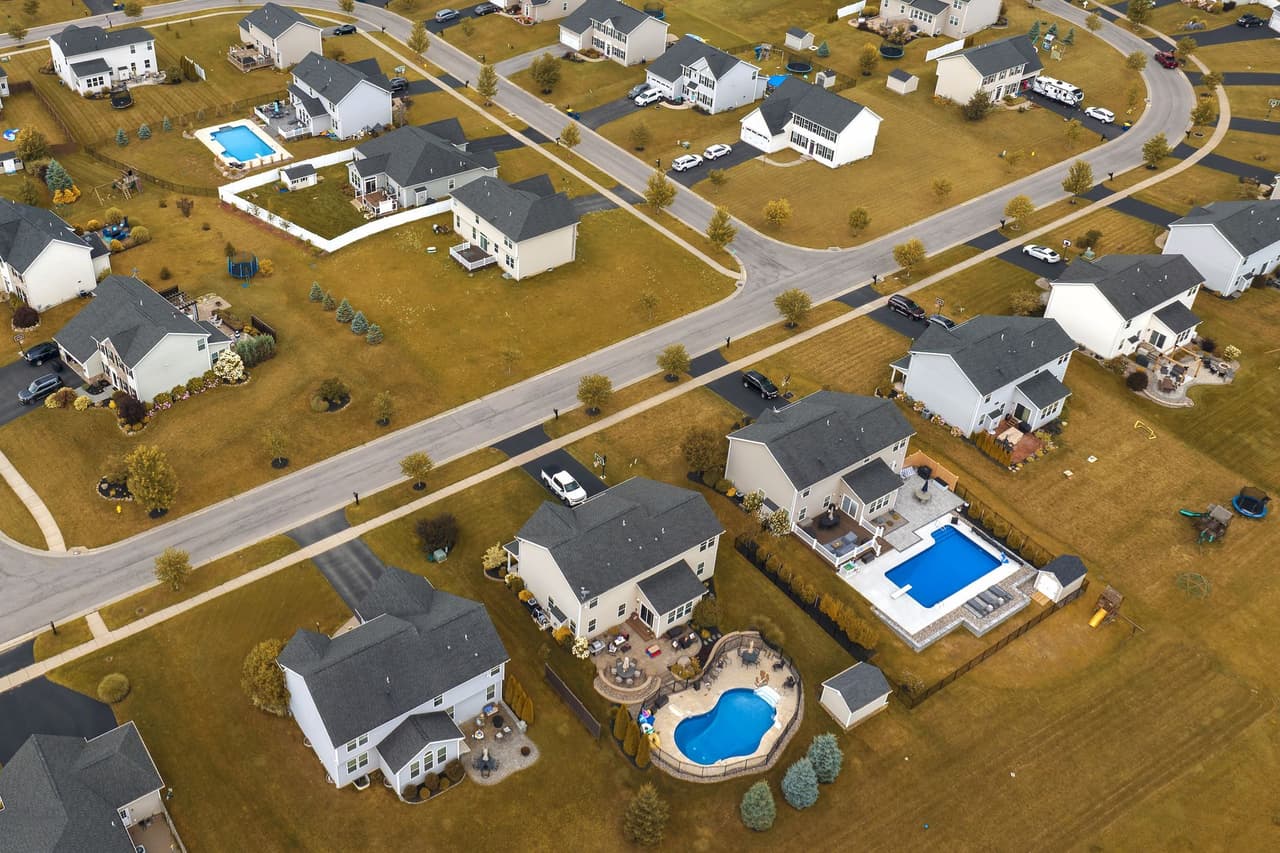 Aerial view of suburban neighborhood with multiple houses, some featuring swimming pools, and intersecting roads.