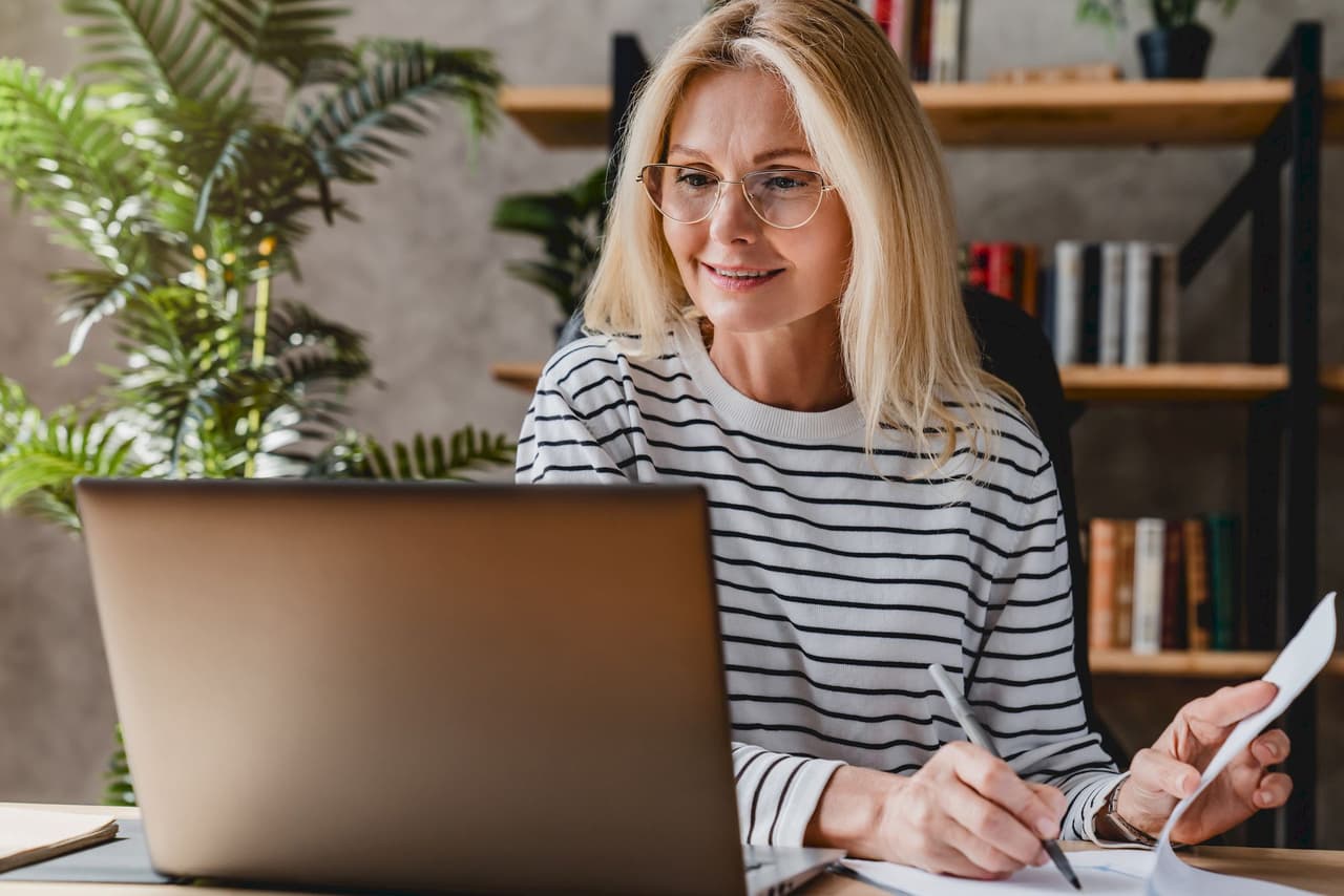 Blonde woman wearing glasses working on a laptop at home with a striped shirt, holding a pen and paper, with a blurred background of bookshelves and plants.