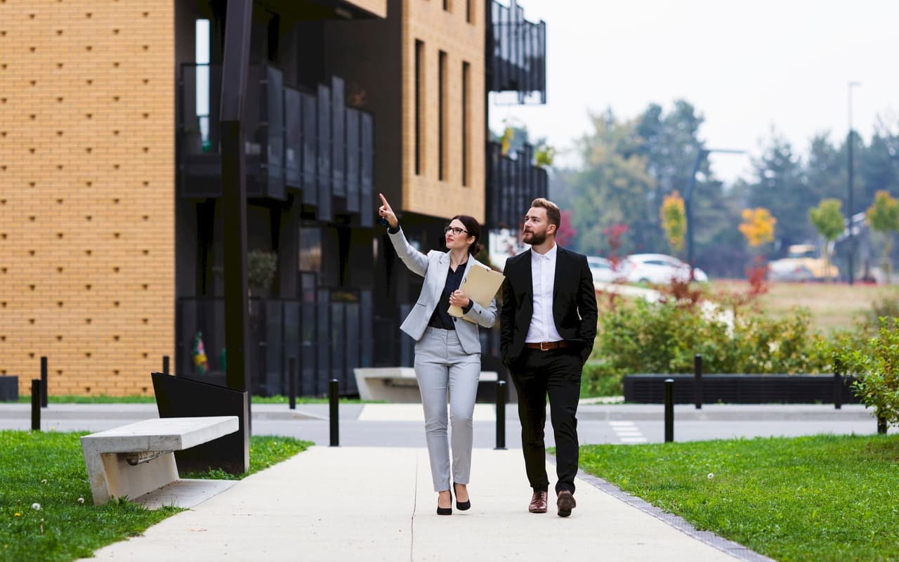 A man and a woman in business attire walking outdoors near modern buildings, engaged in discussion with the woman pointing ahead.