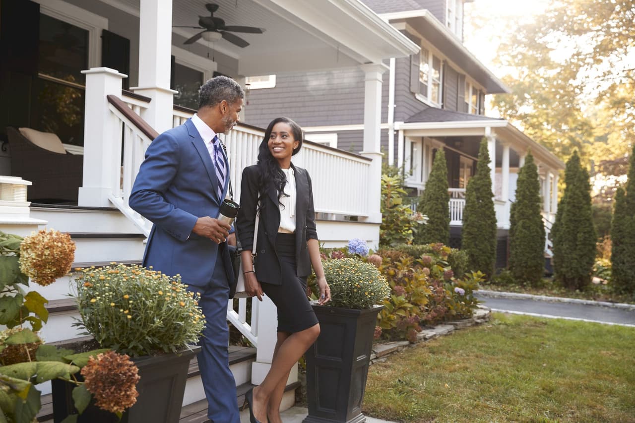 A businessman and a businesswoman walking outdoors near a house porch, engaged in conversation on a sunny day.