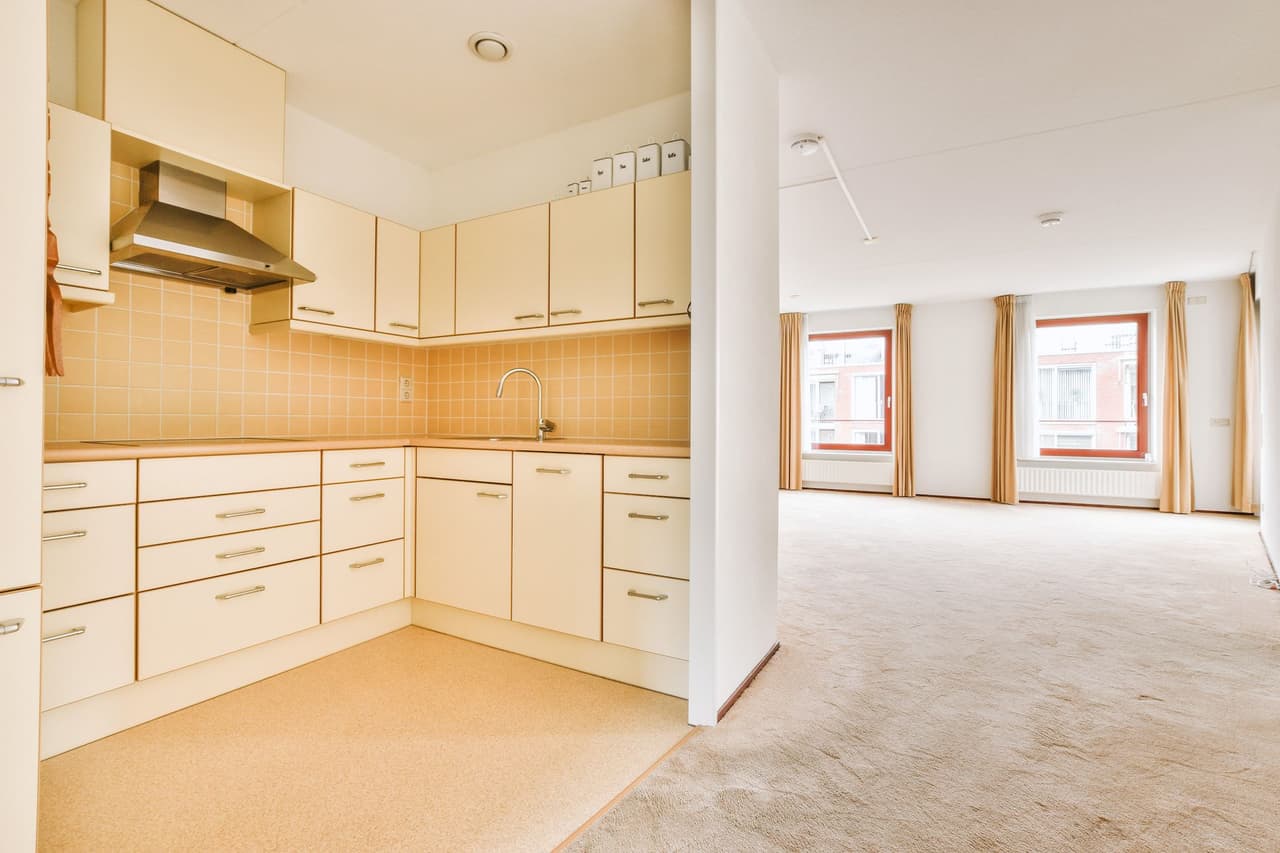 Empty modern kitchen with beige cabinets and tiled backsplash adjacent to a spacious living room with large windows and beige carpet