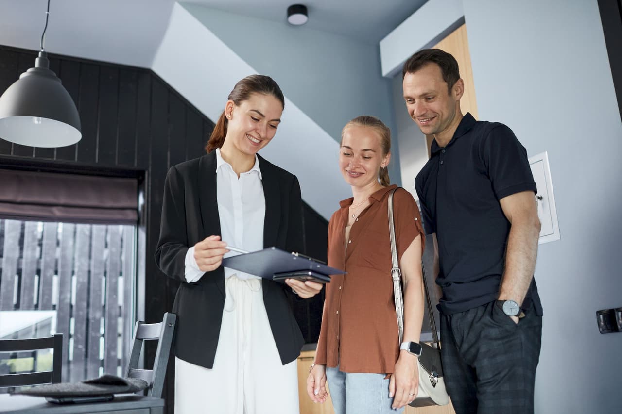 Female real estate agent showing documents to a smiling couple inside a modern home with a black and gray interior