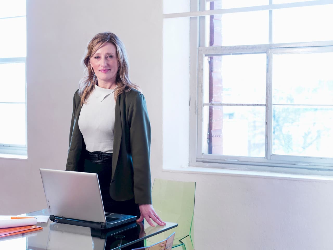 Portrait of a professional woman standing in an office environment with a laptop on a glass table and large windows in the background.