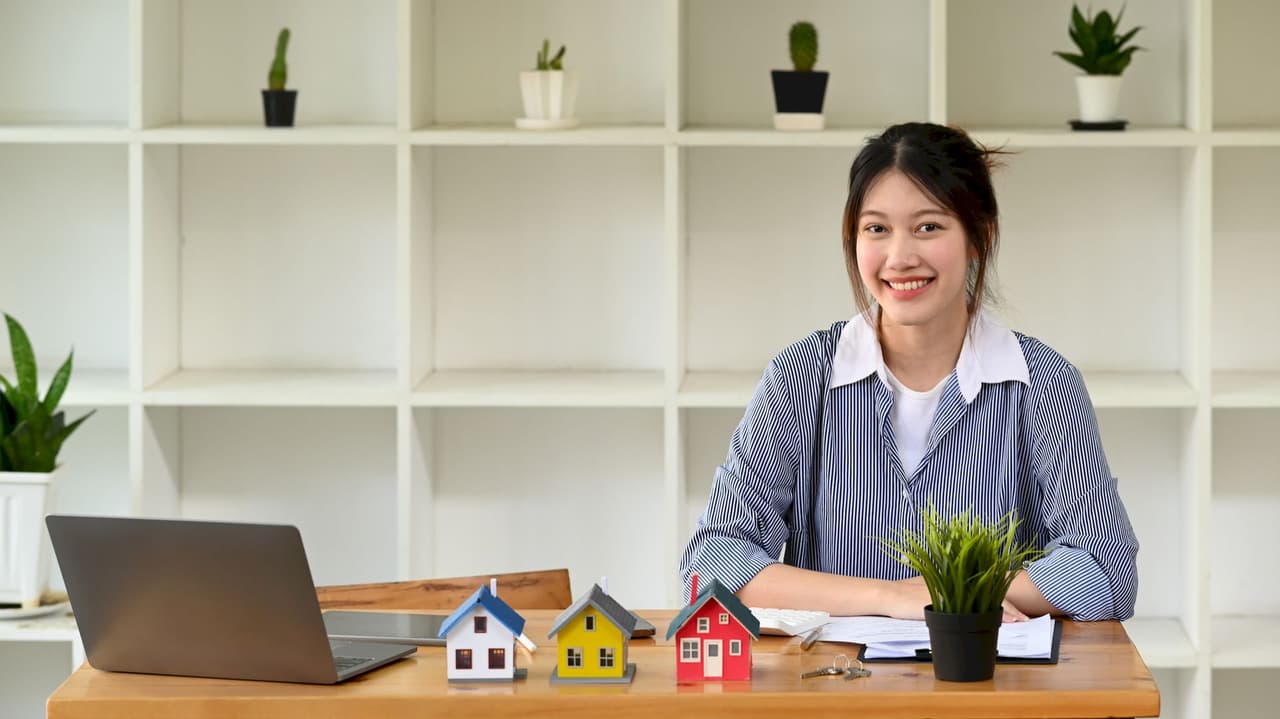 A young female real estate agent sitting at a desk with a laptop, small model houses, and a potted plant, smiling with a white shelf background.