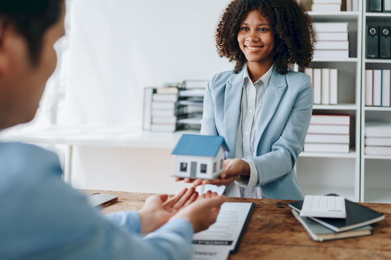 Smiling female real estate agent in a blue blazer holding a small model house, sitting at a desk with documents and notebooks, engaging with a client in an office setting
