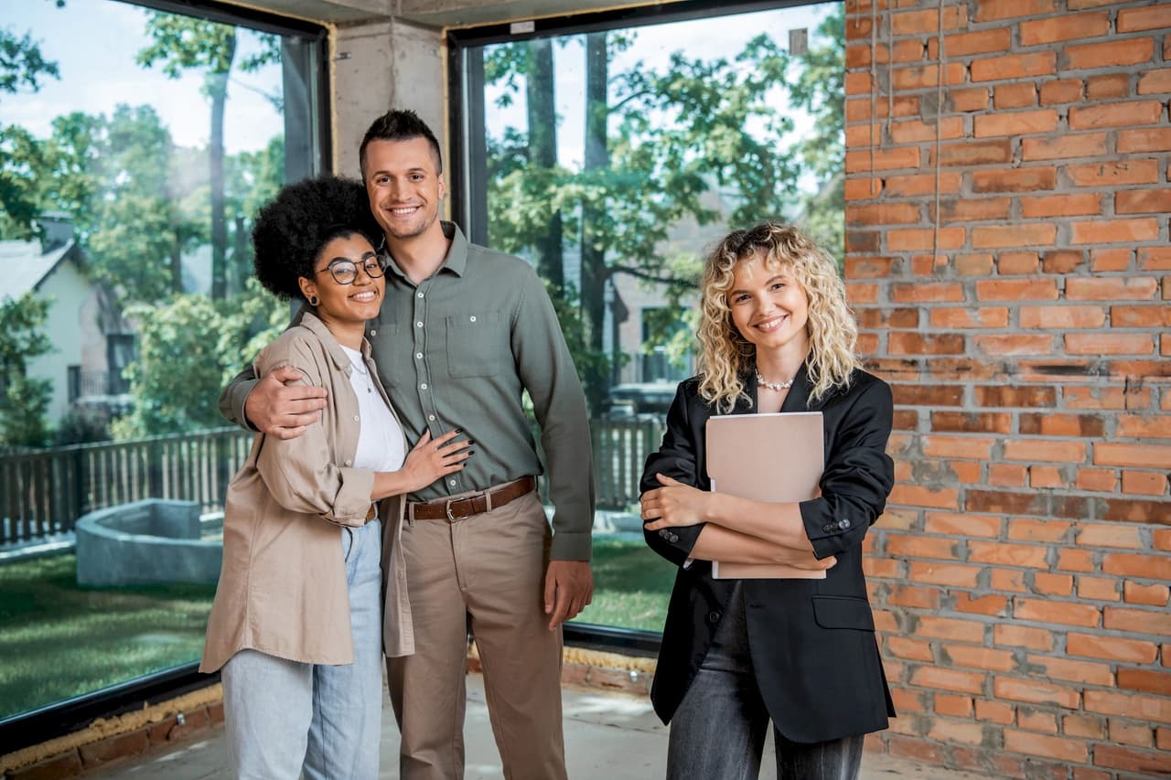 Happy couple standing inside a house with a female real estate agent holding a folder, large windows showing outdoor greenery, and exposed brick wall in the background.