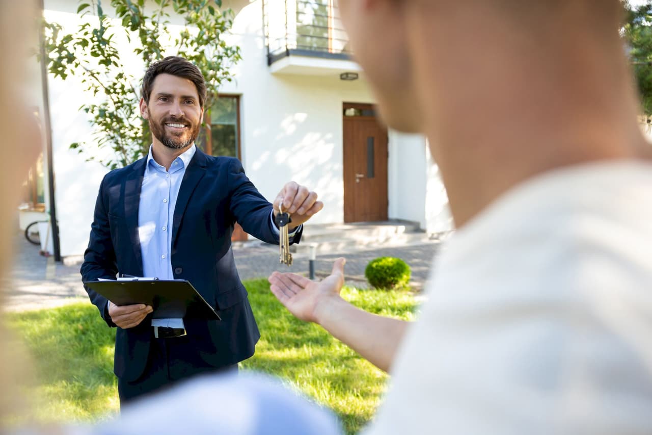 Male real estate agent handing house keys to a person outside a house with a clipboard in hand